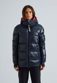 Marinblå pufferjacka med en glänsande, isolerad finish, kontrasterande röd inredning, full dragkedja och huva, med en logotyp på framsidan.