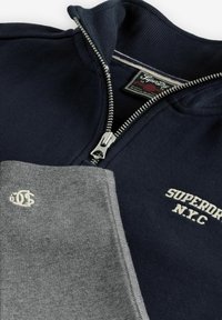 Námornícky zip-up sveter s výšivkou "SUPERDRY N.Y.C" a zloženou šedou tkaninou s malým bielym vyšitým logom.