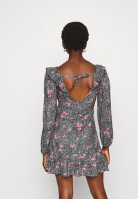 Mujer vestida con un minivestido negro de flores, con mangas largas, dobladillo con volantes y una espalda descubiertaatada con un lazo en los omóplatos.