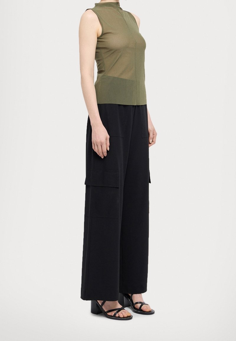 Femme portant un haut sans manches vert olive, un large pantalon cargo noir avec de grandes poches latérales, et des sandales noires à talons cloutées.