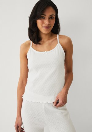 Kvinde med skulderlangt mørkt hår iført en hvid ærmeløs top og matchende bukser med diskrete hjerte-mønstre, stående foran en ensfarvet baggrund.