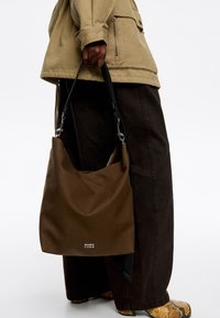 Brązowa miękka torba na ramię z czarnym paskiem, z logo na dole, trzymana przez osobę w casualowym, ciemnym ubraniu.