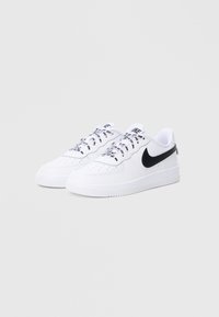 Biele tenisky Nike Air Force 1 majú perforovanú špičku, čierne logo Swoosh na boku a ploché šnúrky s dekoratívnymi vzormi.