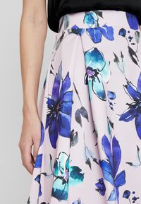 Mano appoggiata accanto a una gonna plissettata con grandi motivi floreali acquerello blu e turchese su tessuto rosa pallido.