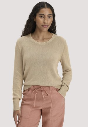 Vrouw met lang krullend haar, die een beige gebreide trui en stoffige roze broek met koord draagt, staand met één hand in de zak.