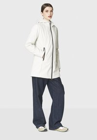 Nainen, jolla on päällä valkoinen huppari vetoketjulla, tummat leveät housut ja valkoiset trainers-kengät, seisoo käsi hupun taskussa.