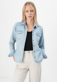 Light blue denim shirt with button closure, two chest pockets, and folded cuffs. Worn over a black top and paired with beige pants.