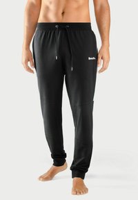 Schwarze Sweatpants mit elastischem Bund und Kordelzug, ausgestattet mit seitlichen Einsätzen und einem gedruckten Logo auf der Vorderseite. Glatte Textur.