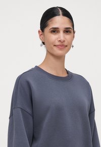 Jauna sieviete ar tumšiem matiem, kas veidojusi zemu bun, valkā sudraba ziedu auskari un brīvu tumši pelēku kreklu ar apaļu kaklu uz neuzkrītoša fona.