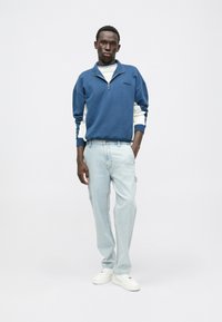 Sudadera azul con cremallera y acentos en las mangas en blanco, combinada con jeans de mezclilla azul claro y zapatillas blancas. Diseño simple y de corte relajado.