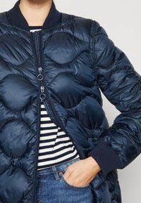 Námořnický puffer kabát s prošívaným vzorem, se zipem vpředu, žebrovanými manžetami a límcem, nošený přes pruhované tričko, s rukou v kapse.