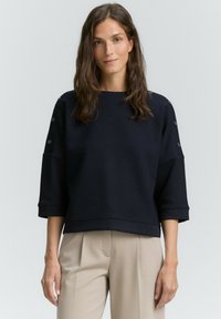 Marineblauwe sweatshirt met textuur, driekwartmouwen, geribde zoom en knoopaccenten op de schouders. Gecombineerd met lichtbeige broeken.