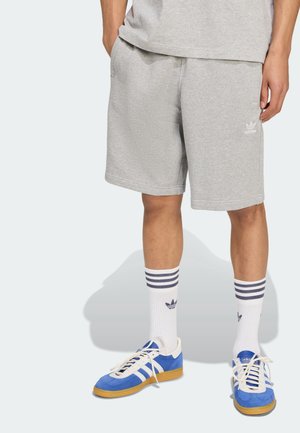 Personne portant un short gris Adidas, des chaussettes blanches Adidas avec des rayures noires, et des baskets bleues Adidas avec des rayures blanches et des semelles en caoutchouc.