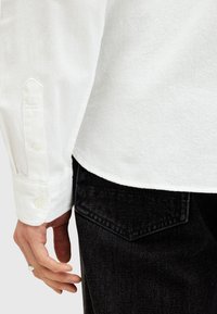Hvid textured skjorte med knapmanchetter, vist sammen med sorte denim jeans med en skrå baglomme. Nærbillede med hånd placeret.