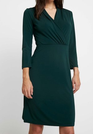 Kobieta w ciemnozielonej sukience do kolan z rękawami do połowy przedramienia i kopertowym dekoltem w kształcie litery V na jednolitym tle.