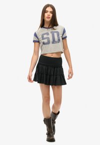 T-shirt gris court avec imprimé "SD" et manches bleues, associée à une mini-jupe noire à volants avec une ceinture froncée et des bottines marron.