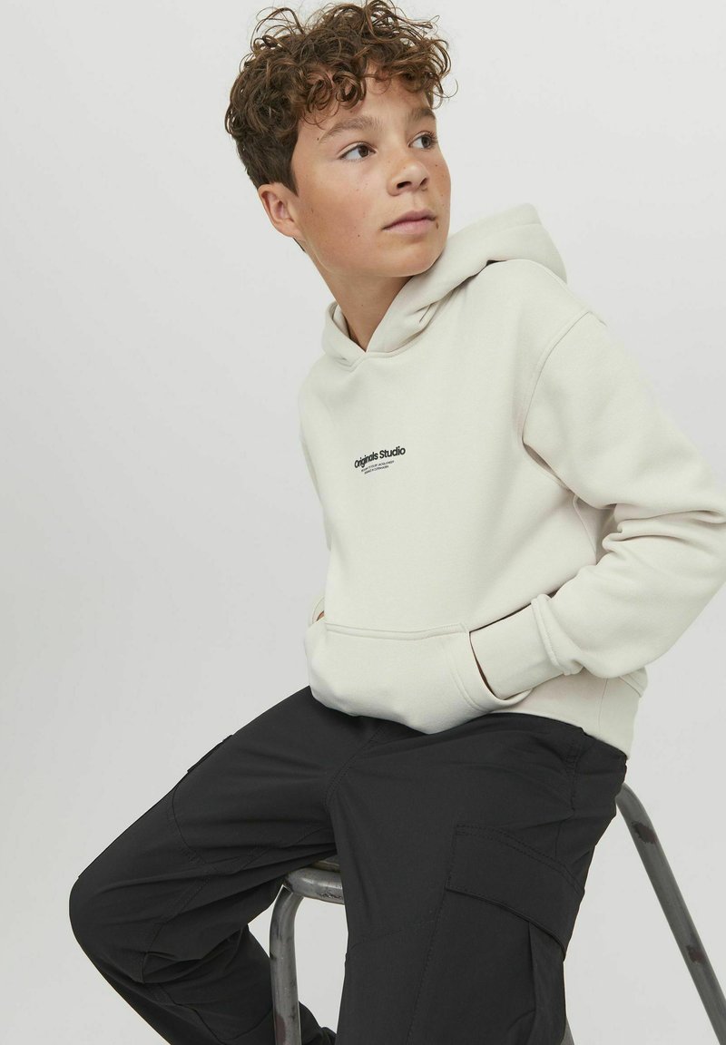 Haut à capuche beige avec une poche avant et un logo noir, associé à un pantalon cargo noir. Tissu doux et texturé, coupe décontractée.