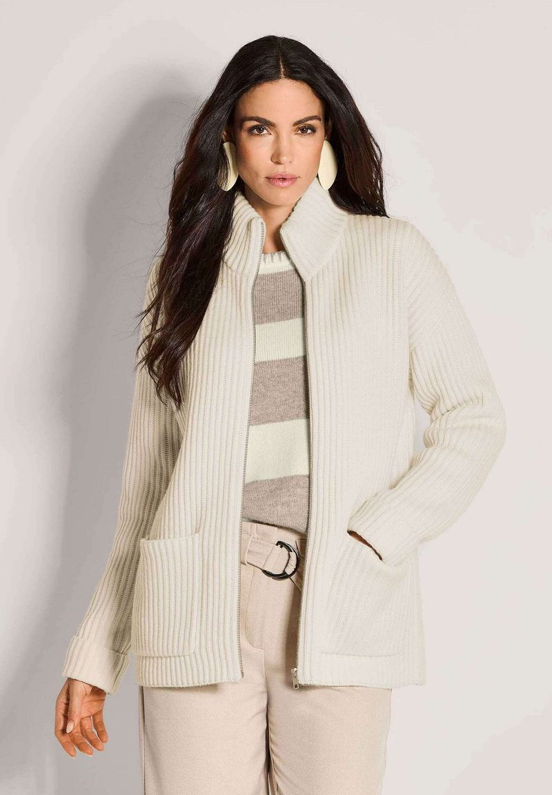 Frau mit langen dunklen Haaren, die große weiße Ohrringe, eine weiße Reißverschluss-Rippstrickjacke, einen gestreiften beige-weißen Pullover und beige Hose trägt.