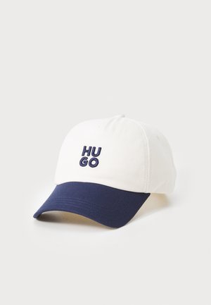 Valkoinen baseball-lippis, jossa on tummansininen lippa, etuosassa tummansinisellä brodeerattu "HU GO" -logo. Materiaali näyttää olevan puuvillaa.