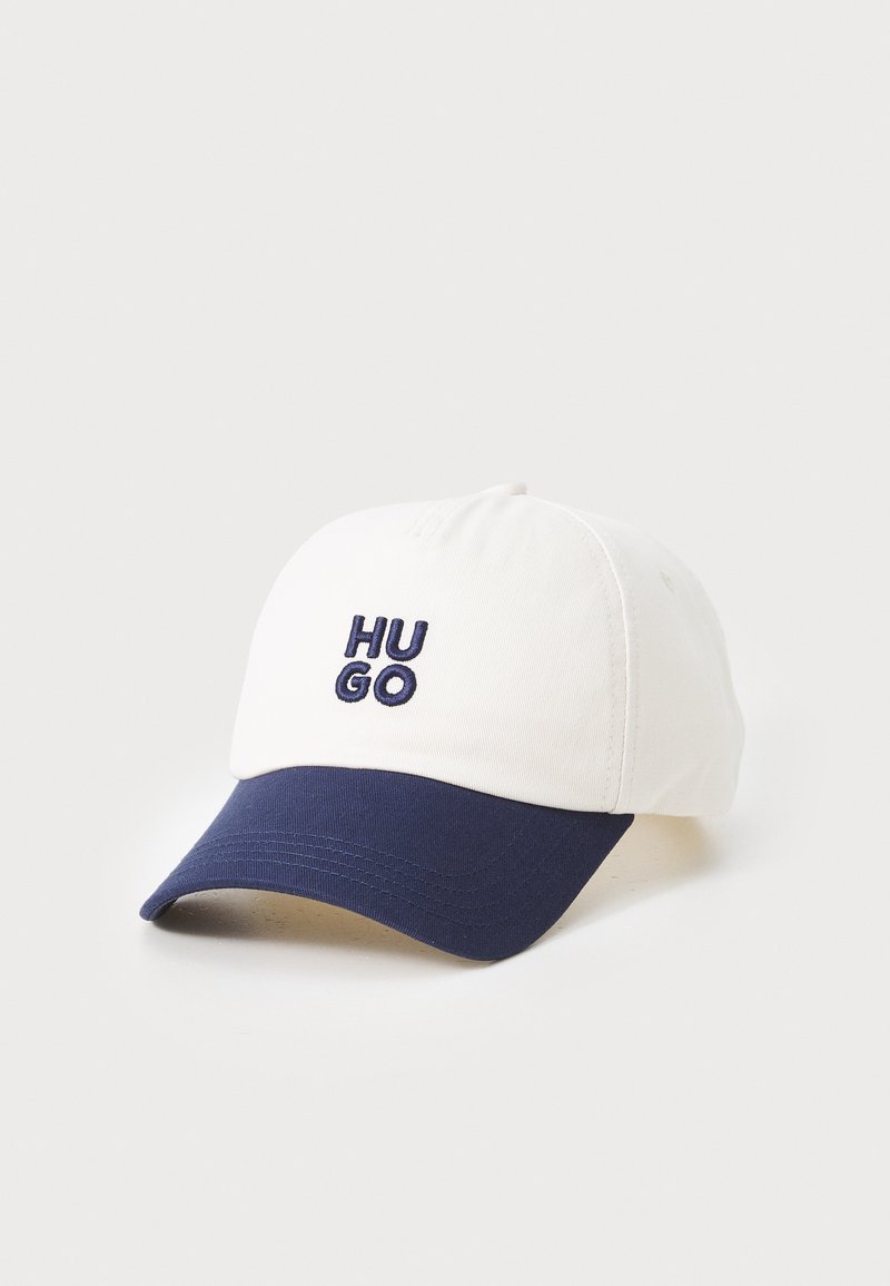 Valge pesapalli müts tumesinise äärega, mille ees on tumedast sinisest tikandiga "HU GO" logo. Materjal näib olevat puuvill.