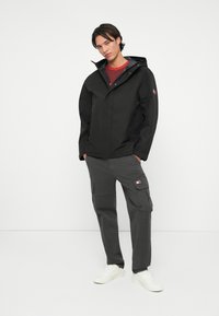 Μαύρο αδιάβροχο μπουφάν με κουκούλα, συνδυασμένο με γκρι παντελόνι cargo που διαθέτει τσέπες και λογότυπο, φορεμένο με λευκά αθλητικά παπούτσια.