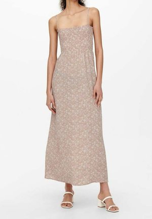 Vrouw draagt een mouwloze, lichtroze maxi-jurk met bloemenprint, dunne bandjes en witte sandalen met blokhakken, staand tegen een witte achtergrond.