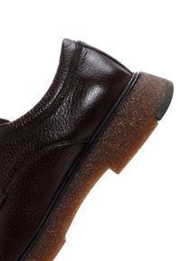 Chaussure en cuir marron foncé avec surface texturée et semelle en caoutchouc marron épaisse avec des crampons, vue sur le côté sur fond blanc.