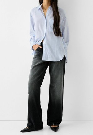 Femme portant un jean noir ample, une chemise à rayures bleues et blanches boutonnée, et des chaussures noires pointues avec clous.