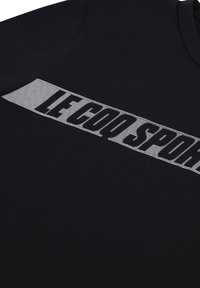 T-shirt en coton noir avec un graphique à rayures blanches horizontales portant l'inscription "LE COQ SPORTIF," avec un col ras du cou et une texture lisse.