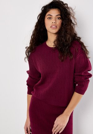 Burgundy knitted sweater with ribbed texture and long sleeves, paired with a matching ribbed skirt, against a neutral background.
