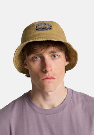 Sombrero tipo bucket en color caqui con un diseño bordado en azul y negro. Tela suave y texturizada con un ala ancha. Llevado con una camiseta de color púrpura claro.