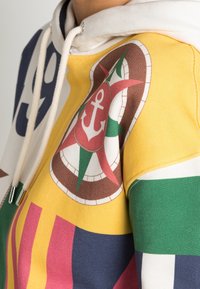 Zbliżenie multikolorowego hoodie z żółtymi, zielonymi i różowymi panelami oraz wzorem kompasu z kotwicą na obszarze klatki piersiowej.