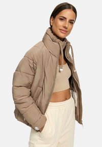 Khaki pufferjack met hoge kraag, ritsluiting en trekkoorden. Beschikt over een gestikte textuur en zijzakken, gelaagd over een beige cropped top.