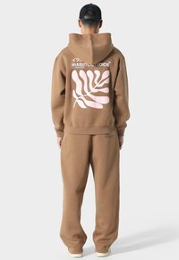 Brauner Kapuzensweater und passende Jogginghose. Enthält ein pinkes botanisches Design auf der Rückseite und gerippte Bündchen an den Handgelenken und am Bund.