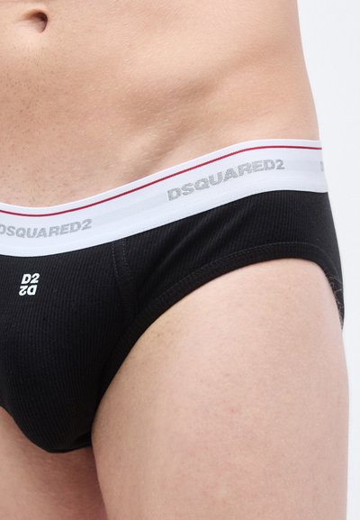 Slip noir côtelé avec une ceinture blanche à accents rouges, logo en relief et coupe ajustée. Détails du tissu texturé visibles.