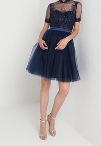 Abito blu navy con corpetto intricatamente decorato con perline e gonna corta in tulle. Maniche trasparenti e vita coordinata. Sandali con tacco argento.
