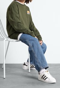 Felpa verde scuro con logo, jeans a gamba larga in denim chiaro e sneakers bianche con strisce nere, seduto su una sedia in metallo bianco.