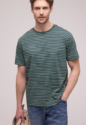 Mężczyzna w zielono-szarej pasiastce z krótkim rękawem i niebieskich dżinsach, trzymający beżowy kapelusz w lewej ręce, na jednolitym tle.