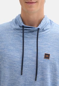 Pullover azzurro chiaro con colletto alto, due lacci neri e un'etichetta quadrata con sfondo nero e un emblema arancione.