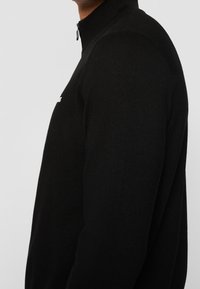 Black zip-up sweater featuring a high collar, ribbed shoulder detailing, and a smooth texture. Fabric appears soft and form-fitting.