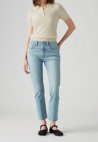 Polo de color amarillo claro combinado con jeans ajustados de color azul claro. Los jeans tienen un diseño clásico de cinco bolsillos y un dobladillo deshilachado. Unos zapatos planos negros completan el look.