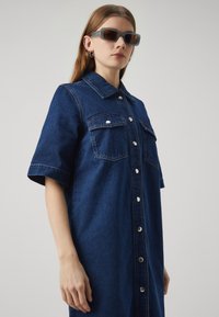 Robe chemise en denim bleu foncé, avec des manches courtes, un bouton devant, deux poches poitrine et des boutons en métal argenté. Texture lisse.