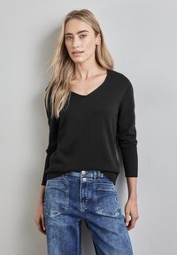 Schwarzer V-Ausschnitt-Pullover mit langen Ärmeln, kombiniert mit hellblauen Jeans. Der Pullover hat einen gerippten Saum und Ärmelabschlüsse.