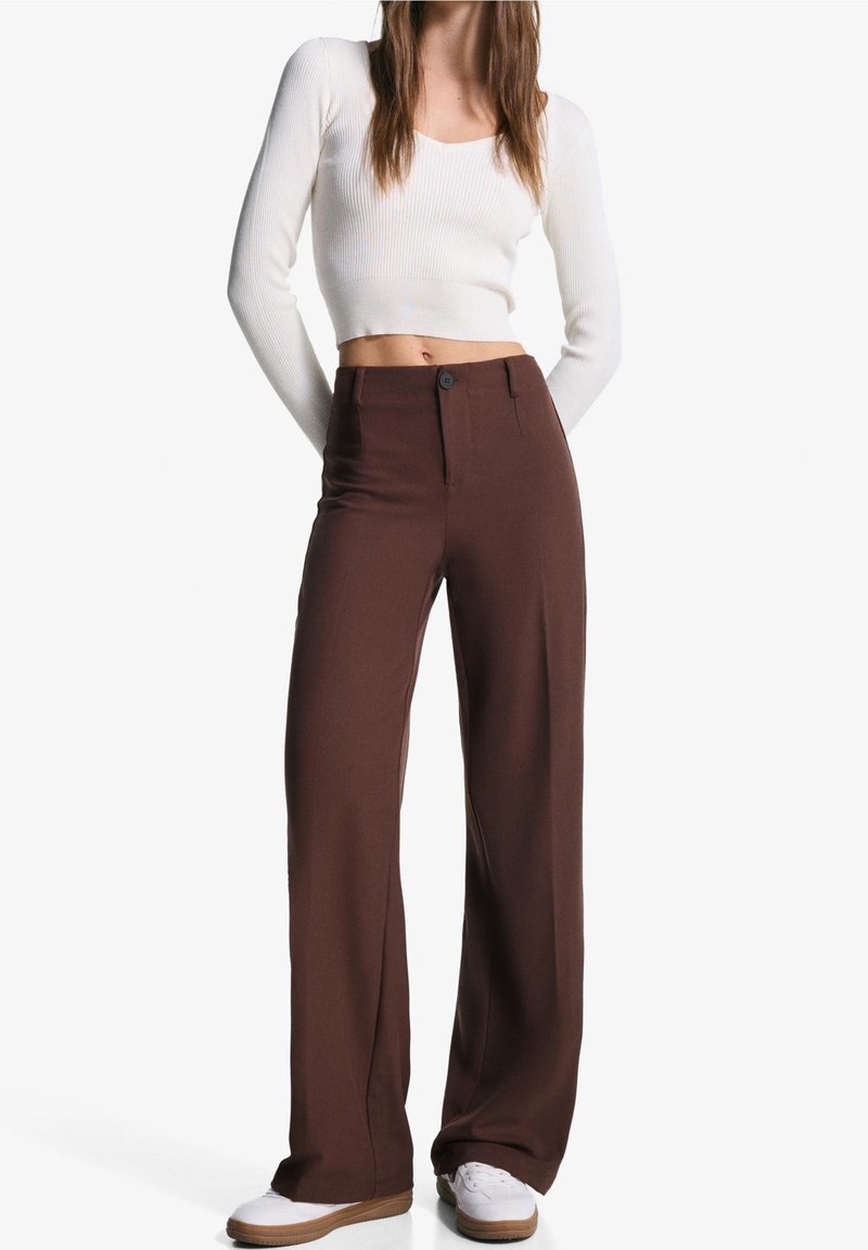 Femme portant un crop top blanc à manches longues, un pantalon marron taille haute à jambes larges et des baskets blanches, debout les mains derrière le dos.