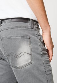 Jeans de mezclilla gris con un acabado desgastado, que cuentan con un diseño de bolsillo trasero de dos patrones zigzagueantes superpuestos y una etiqueta de cuero negra en la cintura.