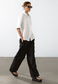 Witte linnen blouse met knopen, zwarte wijde broekspijpen en chunky zwarte platformsandalen. Accessoires omvatten een zilveren armband en een zonnebril.