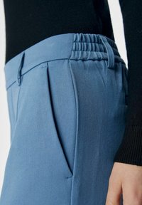 Lys blå skræddersyede bukser med et struktureret stof, plisseret fortil og elastisk talje. Sort sweaterærme synligt i baggrunden.