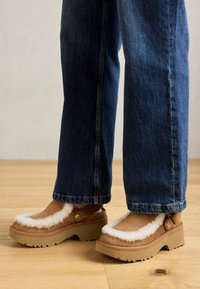 Bruine leren klompen van suède met een witte nepbont rand en tan rubberen zolen, uitgerust met zijgespen en een chunky zool, gedragen met blauwe jeans.