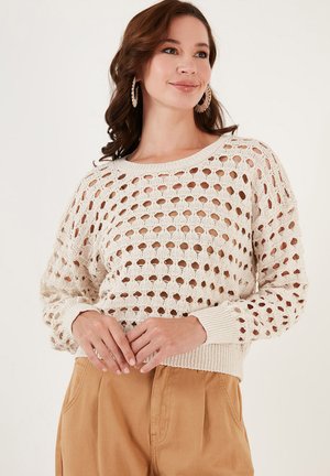 Crème-kleurige gebreide trui met een groot open-holes ontwerp, lange mouwen en ribbels aan de manchetten en onderkant. Draag het met lichtbruine high-waisted broeken.