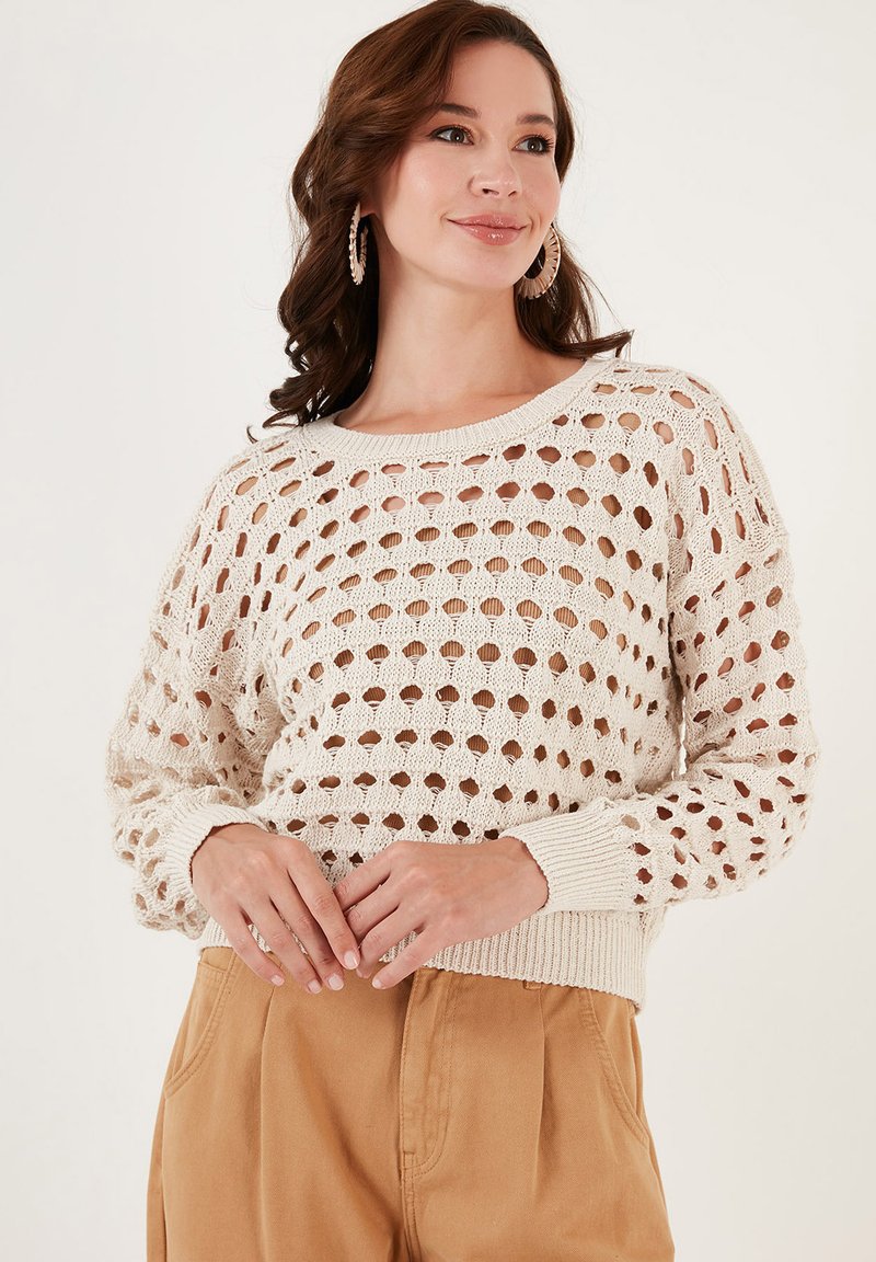 Cremefarvet strikket sweater med et stort hulmønster, lange ærmer og ribbede manchetter og kant. Bæres med lysebrune højtaljede bukser.